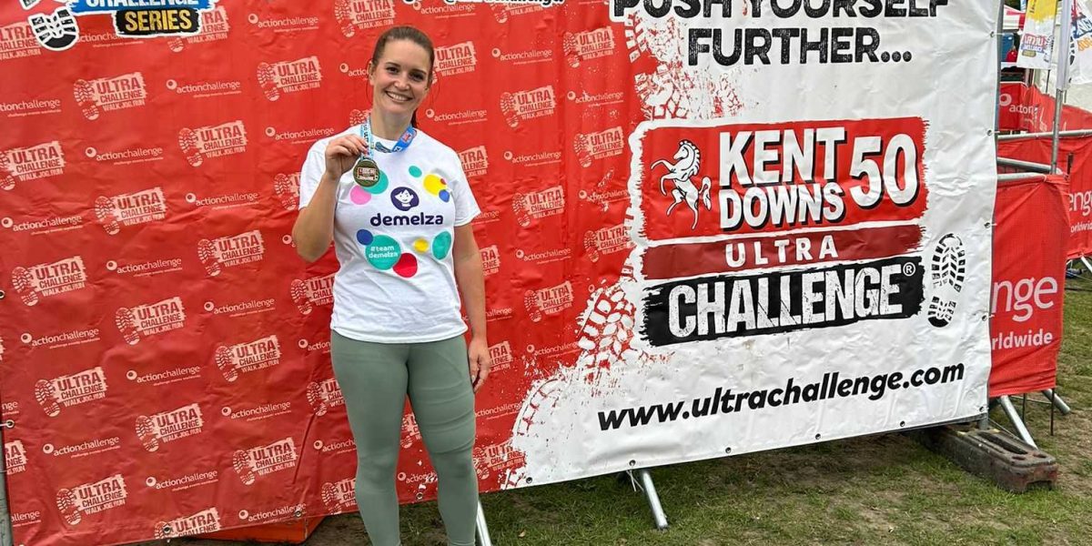 Comparison Tech colleague Dani poses with her medal in front of a sign for the Kent Downs 50 Ultra Challenge.