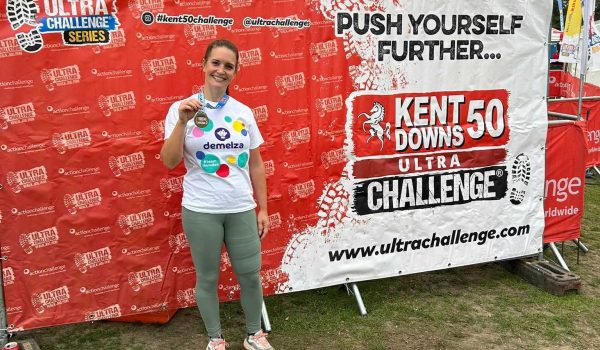 Comparison Tech colleague Dani poses with her medal in front of a sign for the Kent Downs 50 Ultra Challenge.